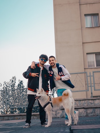 Two young men are posing casually outdoors, standing together with a friendly dog. They are wearing casual streetwear, including jackets and sneakers. The background features a building and some greenery, suggesting an urban environment.