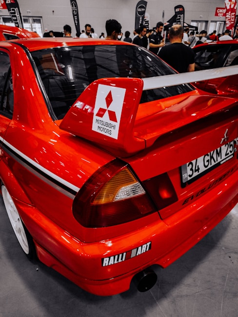 A bright red Mitsubishi car with a large rear spoiler and Ralliart branding is on display indoors. Several people are mingling and observing the surroundings in a spacious exhibition hall, with banners visible in the background.