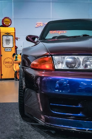 A sleek, shiny car with a metallic finish is prominently displayed in the foreground. The car is dark-colored with hints of purple and blue, and it features a stylish front light and distinctive curves. In the background, there is a vintage Shell gasoline pump with a bright yellow and red color scheme. Neon signage with cursive writing is also visible behind the car.