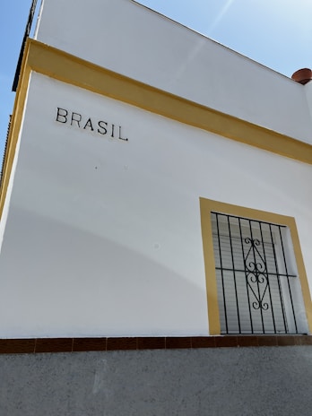 A corner of a white building with yellow trim is visible. The word 'BRASIL' is written on the wall. There is a window with a black decorative wrought iron grill on the right side, framed in yellow.