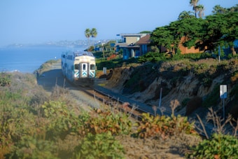 A train travels along a coastal railway track, bordered by lush greenery and residential houses. The ocean is visible on one side, with gentle waves creating a serene seascape. Palm trees and vibrant vegetation add to the scenic landscape, while the train follows the winding rail path.