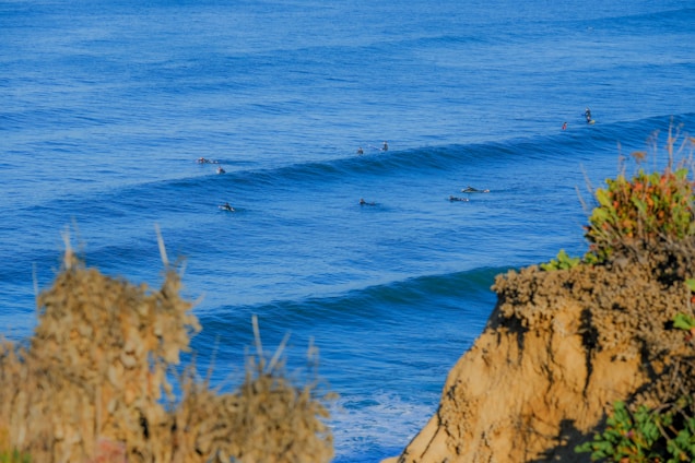 Several surfers are floating on their boards in the ocean, waiting for waves. The ocean is a vibrant blue, and the waves are gently rolling. In the foreground, there are rocky cliffs with some sparse vegetation.