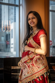 A woman dressed in a vibrant red traditional outfit with intricate gold and green embroidery stands indoors beside large windows. Her long black hair cascades down her shoulders, and she is smiling warmly while cradling her stomach, suggesting that she might be pregnant. The background features an urban exterior view with tall buildings and reflective surfaces.