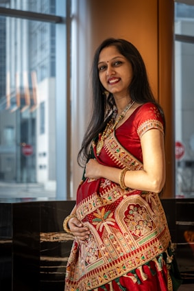 A woman dressed in a vibrant red traditional outfit with intricate gold and green embroidery stands indoors beside large windows. Her long black hair cascades down her shoulders, and she is smiling warmly while cradling her stomach, suggesting that she might be pregnant. The background features an urban exterior view with tall buildings and reflective surfaces.