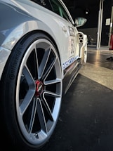 A close-up view of a car with a focus on the wheel and side panel. The wheel features a sleek, modern design with a striking red center cap marked 'rotiform.' The car is decorated with decals, including a racing number '53' and a blue stripe, indicating it's likely used for racing or a car event. Background lighting highlights the car's contours, accentuating its sporty appearance.