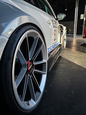 A close-up view of a car with a focus on the wheel and side panel. The wheel features a sleek, modern design with a striking red center cap marked 'rotiform.' The car is decorated with decals, including a racing number '53' and a blue stripe, indicating it's likely used for racing or a car event. Background lighting highlights the car's contours, accentuating its sporty appearance.