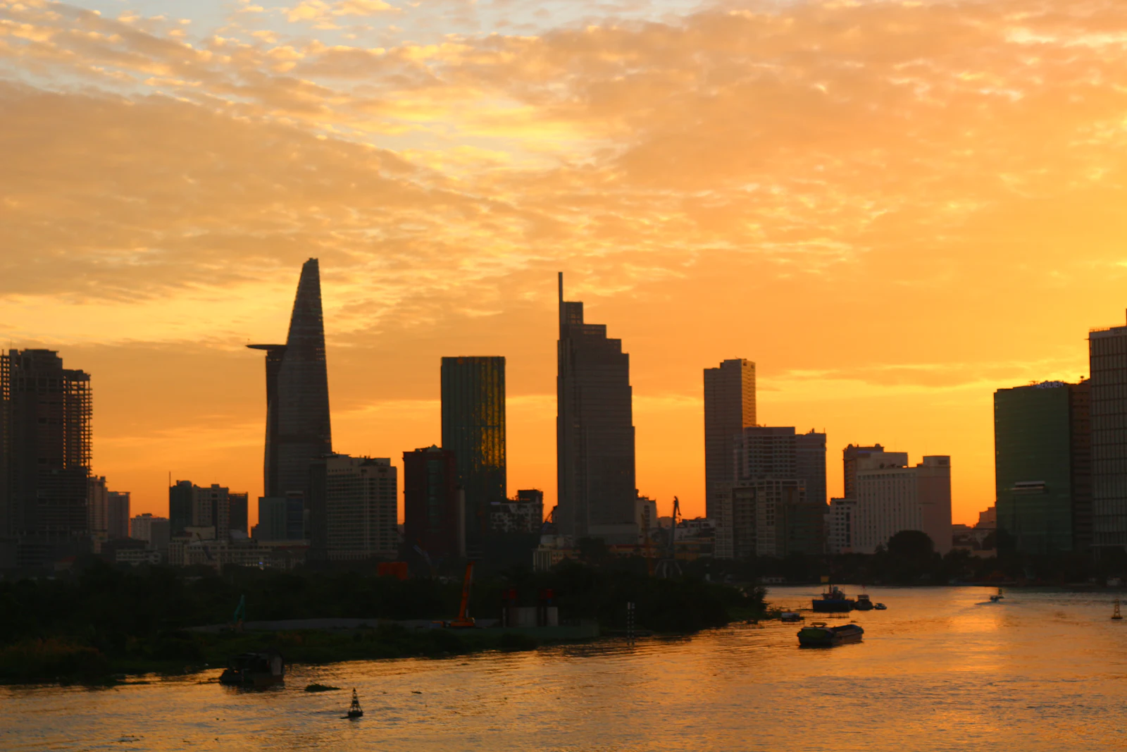 The Ho Chi Minh City skyline silhouetted against a golden sunset over the Saigon River with boats on the water