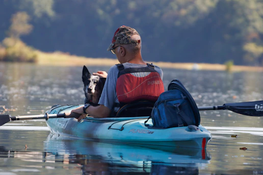 A man wearing a camouflage cap, red life jacket, and gray shirt is paddling a kayak on a serene lake. Next to him in the kayak is a black and white dog. The background features blurred trees and a gently reflective water surface, creating a peaceful and quiet atmosphere.