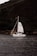 A sailboat with white sails navigates through a calm body of water, with a lush green hillside in the background. Several people are visible on the boat, engaging in various activities. The hillside is dense with trees and vegetation, creating a natural, serene backdrop.