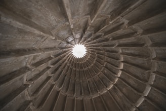 A spiraling concrete structure with ribbed sections, converging towards a bright opening at the top center, suggesting a tunnel or architectural feature with an industrial aesthetic.