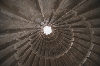 A spiraling concrete structure with ribbed sections, converging towards a bright opening at the top center, suggesting a tunnel or architectural feature with an industrial aesthetic.