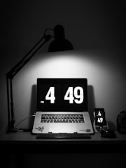 A dimly lit workspace features a laptop displaying a digital clock showing the time 4:49 PM. Nearby, a smartphone mirrors the same time. The workspace is primarily black with a stylish desk lamp casting light directly on the laptop.