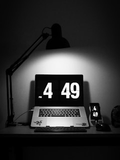 A dimly lit workspace features a laptop displaying a digital clock showing the time 4:49 PM. Nearby, a smartphone mirrors the same time. The workspace is primarily black with a stylish desk lamp casting light directly on the laptop.