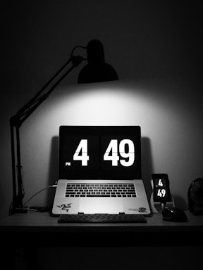 A dimly lit workspace features a laptop displaying a digital clock showing the time 4:49 PM. Nearby, a smartphone mirrors the same time. The workspace is primarily black with a stylish desk lamp casting light directly on the laptop.