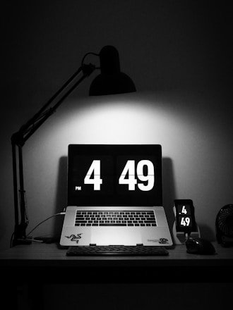 A dimly lit workspace features a laptop displaying a digital clock showing the time 4:49 PM. Nearby, a smartphone mirrors the same time. The workspace is primarily black with a stylish desk lamp casting light directly on the laptop.