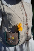 A knitted beige cardigan with wooden buttons and a detailed close-up of a pocket. A bright yellow flower accents the pocket, which is decorated with colorful embroidered flowers. The clothing features a white blouse with a scalloped collar underneath.