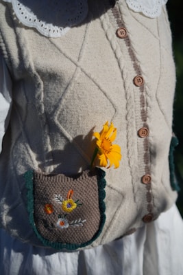 A knitted beige cardigan with wooden buttons and a detailed close-up of a pocket. A bright yellow flower accents the pocket, which is decorated with colorful embroidered flowers. The clothing features a white blouse with a scalloped collar underneath.