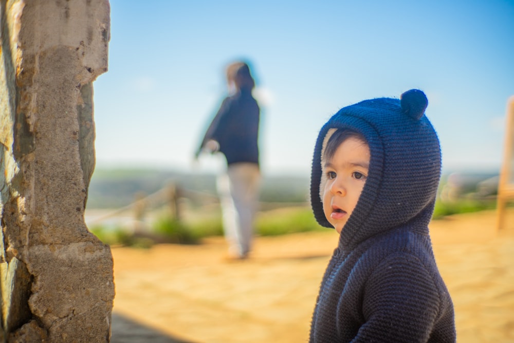 a little boy in a blue sweater and a woman walking in the background