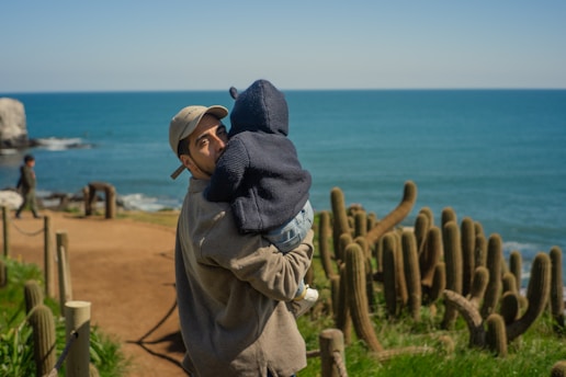 A man wearing a cap and casual clothing is holding a baby wrapped in a dark hooded jacket. They are standing on a path alongside a landscape filled with cactus plants. In the background, there is a view of the ocean under a clear blue sky. Another person is seen walking or standing farther along the path.
