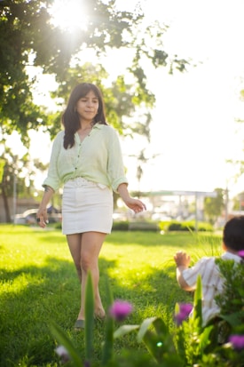 A woman is walking through a sunlit park with trees in the background. She is wearing a light green blouse and a white skirt, and appears calm and content. In the foreground, there are green plants and pink flowers. A child is visible from behind, sitting or crouching on the grass.