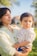 A woman gently holds a young child in an outdoor setting with lush greenery. She appears serene with her eyes closed, while the child looks off into the distance thoughtfully. The bright sunlight casts soft shadows, creating a warm and peaceful atmosphere.
