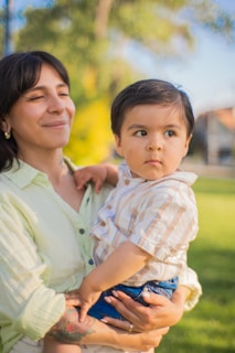 A young woman holding a toddler, both captured in a serene outdoor setting. The woman has short dark hair and is smiling with her eyes closed, while the toddler looks off to the side with a thoughtful expression. The background is a soft focus of greenery and sky.
