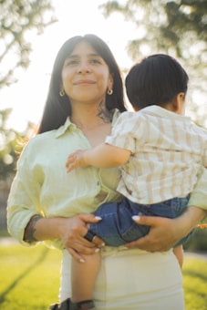 A woman with long dark hair and a tattoo on her neck is holding a small child in her arms outdoors. She wears a light-colored shirt and the child is dressed in a striped shirt and dark jeans. The background is softly blurred, with sunlight filtering through trees, creating a warm glow.