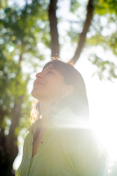 A person is looking upwards with a serene expression, standing in a sunlit forest area. Sunlight creates a glow around them, casting lens flares and enhancing the peaceful atmosphere. The trees are lush, and the background is blurred, creating a bokeh effect.