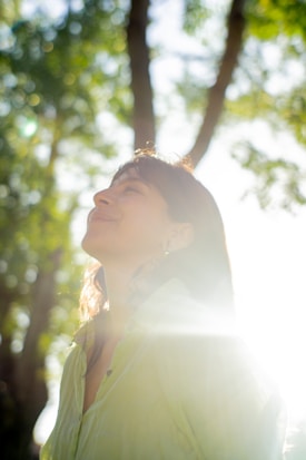 A person is looking upwards with a serene expression, standing in a sunlit forest area. Sunlight creates a glow around them, casting lens flares and enhancing the peaceful atmosphere. The trees are lush, and the background is blurred, creating a bokeh effect.