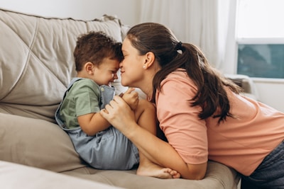 A mother and son share a tender moment on a sofa. The mother, with long dark hair tied back, leans in to touch her forehead against her son's. The child wears a green shirt and light blue overalls, sitting curled up on the sofa with a coy smile. The room has a soft and cozy feel, with light pouring in from a nearby window.