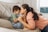 A mother and son share a tender moment on a sofa. The mother, with long dark hair tied back, leans in to touch her forehead against her son's. The child wears a green shirt and light blue overalls, sitting curled up on the sofa with a coy smile. The room has a soft and cozy feel, with light pouring in from a nearby window.