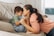 A mother and son share a tender moment on a sofa. The mother, with long dark hair tied back, leans in to touch her forehead against her son's. The child wears a green shirt and light blue overalls, sitting curled up on the sofa with a coy smile. The room has a soft and cozy feel, with light pouring in from a nearby window.