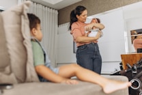 A woman is gently cradling a baby in her arms while standing in a living room setting. A young child sits on a sofa in the foreground, looking towards the woman and baby. The room features neutral tones with white curtains and modern decor.
