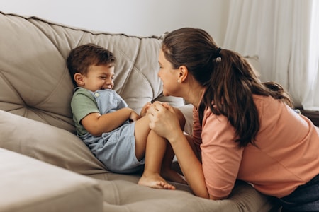 A woman is leaning towards a young boy who is sitting on a couch. The boy is laughing joyfully with his hands covering his face in a playful manner. The woman appears to be engaging warmly with the child, creating an atmosphere of affection and happiness.