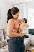 A woman is holding a baby in her arms, looking down at the child with a gentle expression. The woman is wearing a peach-colored shirt and dark jeans, while the baby is wrapped in a light blanket and wearing a pink headband. They are situated in a cozy living room with a sofa and a curtained window in the background.