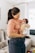 A woman is holding a baby in her arms, looking down at the child with a gentle expression. The woman is wearing a peach-colored shirt and dark jeans, while the baby is wrapped in a light blanket and wearing a pink headband. They are situated in a cozy living room with a sofa and a curtained window in the background.