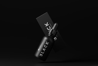A black dropper bottle labeled 'BLACK' is positioned in front of a matching black box on a dark background, giving a sleek and minimalist appearance.