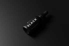 A sleek black dropper bottle with the word 'BLACK' prominently displayed on its label lies against a dark gradient background. The bottle's design is minimalistic and modern, with a matte finish and a circular cap.