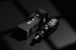 A black bottle labeled 'BLACK' lies next to a matching black box with similar branding in a minimalist, monochromatic setting. The image emphasizes luxury and exclusivity through its use of shadow and light, highlighting the sleek design of the packaging.