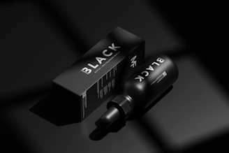 A black bottle labeled 'BLACK' lies next to a matching black box with similar branding in a minimalist, monochromatic setting. The image emphasizes luxury and exclusivity through its use of shadow and light, highlighting the sleek design of the packaging.