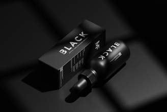 A black bottle labeled 'BLACK' lies next to a matching black box with similar branding in a minimalist, monochromatic setting. The image emphasizes luxury and exclusivity through its use of shadow and light, highlighting the sleek design of the packaging.