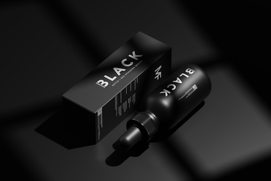 A black bottle labeled 'BLACK' lies next to a matching black box with similar branding in a minimalist, monochromatic setting. The image emphasizes luxury and exclusivity through its use of shadow and light, highlighting the sleek design of the packaging.