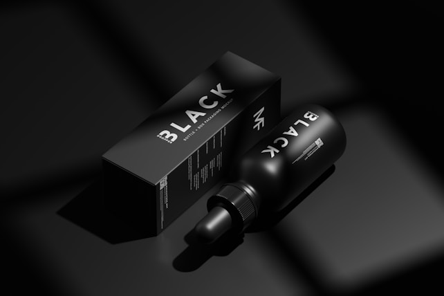 A black bottle labeled 'BLACK' lies next to a matching black box with similar branding in a minimalist, monochromatic setting. The image emphasizes luxury and exclusivity through its use of shadow and light, highlighting the sleek design of the packaging.