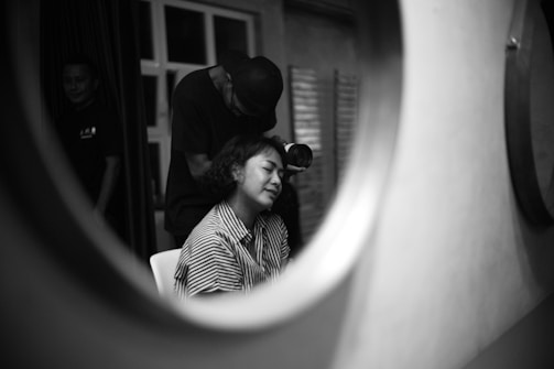 A black and white photo captured through a round reflective surface. It features a woman with closed eyes sitting on a chair while another person, wearing a cap, is attending to her hair with a hairdryer. Another person stands in the background near a window with curtains partially drawn.
