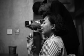 A black and white photograph featuring a woman in the foreground with closed eyes and hands raised slightly, possibly in an expressive or emotional state. Behind her, a man wearing glasses and a cap is holding a camera, pointing it towards her, as if documenting the moment. The setting appears to be indoors with a wall and some objects in the background.