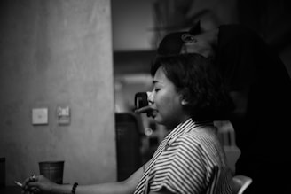A black and white photo capturing a woman in profile, seated with eyes closed, appearing to be in a moment of reflection or deep thought. A person behind her, whose face is not visible, seems to be adjusting her hair or giving a massage. The setting appears to be indoors with a soft focus on the background.