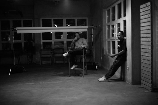 A black and white image featuring two people in a dimly lit room. One person is sitting on a stool, appearing relaxed, while the other leans against a wall near a set of windows. The room has a minimalist and industrial feel, with visible lighting equipment in the scene.