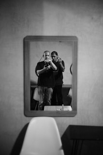 Two individuals are captured in a mirror reflection. One is slightly in front, holding a camera, and the other is behind. They are standing in a room with minimalist decor, featuring a table and chairs. The scene is in black and white, adding a classic feel to the composition.