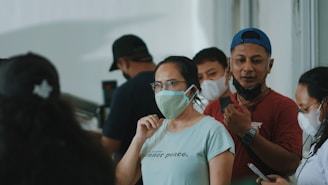 A group of people standing close together indoors, wearing masks. The focus is on a woman in a light green shirt and glasses, who is looking forward with a neutral expression. The people around her are also masked, suggesting adherence to health guidelines.
