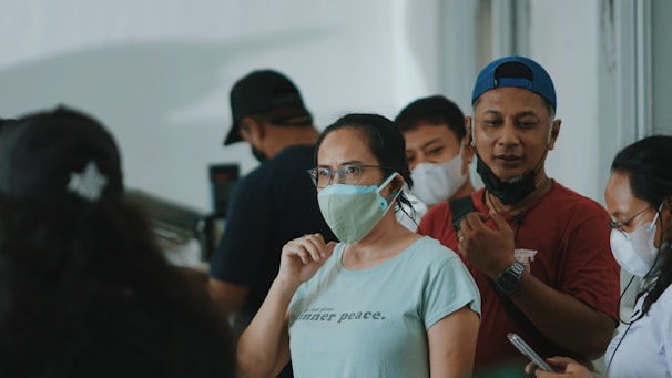 A group of people standing close together indoors, wearing masks. The focus is on a woman in a light green shirt and glasses, who is looking forward with a neutral expression. The people around her are also masked, suggesting adherence to health guidelines.
